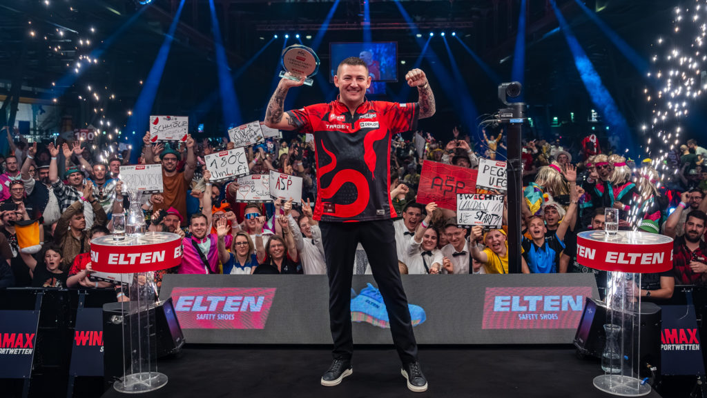 Nathan Aspinall posiert als glücklicher Sieger des ELTEN Safety Shoes German Darts Grand Prix 2026 vor jubelndem Publikum in München